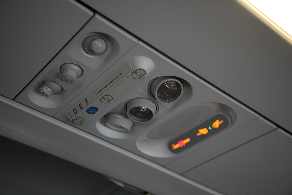 An aircraft passenger-service panel contains air vents, reading lights, seat letters and illuminated signs.