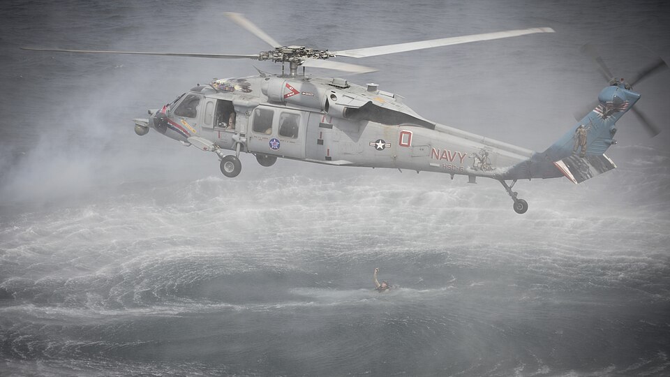 Rescue helicopter hovering above rough water with heavy spray beneath its rotor.