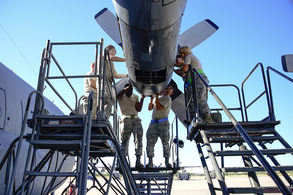 Technicians on work platforms beneath an aircraft engine, surrounded by stairs and handrails.