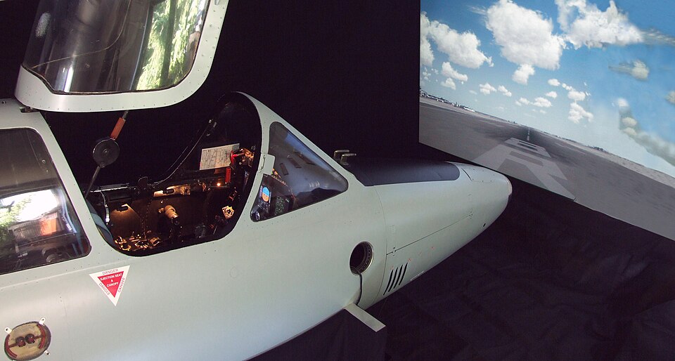 Cockpit simulator with an open canopy facing a projected runway-and-sky display.