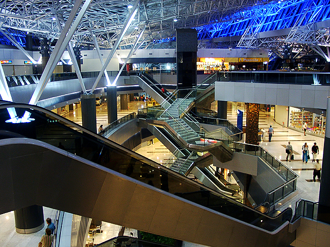 Escalators connect open terminal levels beneath a large steel roof structure and blue lighting.