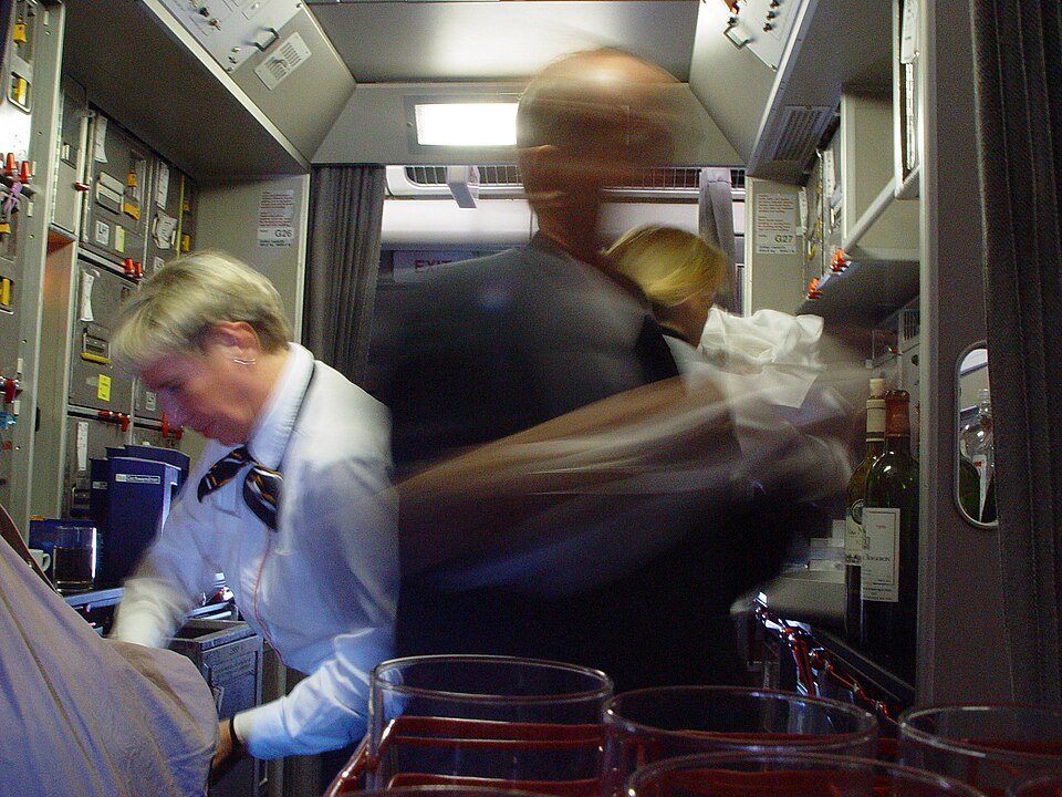 Two cabin crew members work in a narrow aircraft galley, with one blurred by movement.