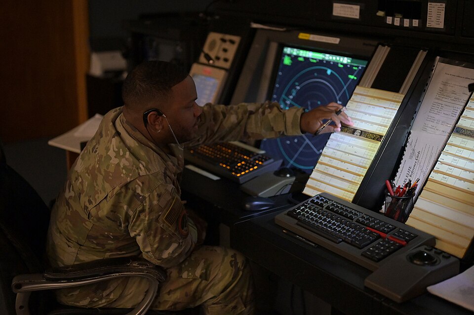 Seated uniformed controller reaching toward a rack beside radar and computer displays.