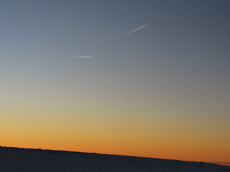 Several pale linear contrails above a dark ridge and orange sunset horizon.