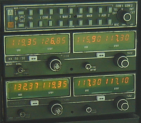 Stacked aircraft radio panels with green frequency displays, buttons and tuning knobs.