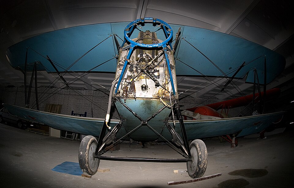 Front view of a historic biplane with exposed metal framework, landing wheels and bracing wires.