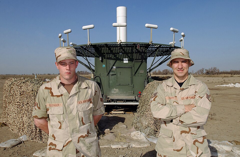 Two uniformed technicians in front of a raised circular antenna ring and green equipment shelter.