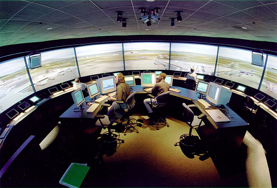 Wide curved control-room consoles facing panoramic simulated airport scenery.