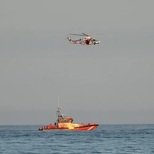 A red-and-white helicopter hovering above an orange vessel at sea.