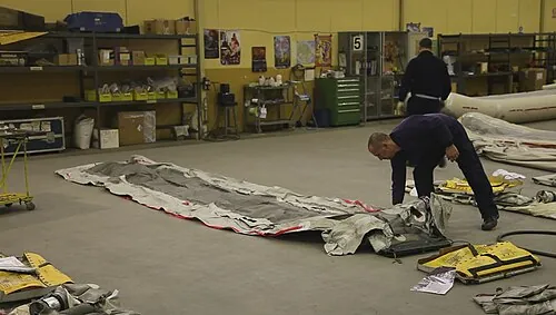 People working beside uninflated evacuation-slide material laid out on a workshop floor.