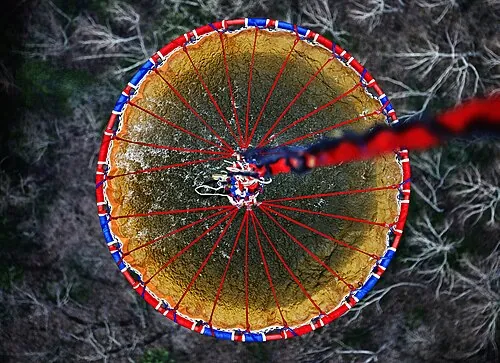Downward view of a water-filled firefighting bucket suspended on a cable above trees.