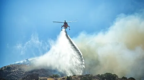 A helicopter releasing a broad stream of water over a smoky hillside.