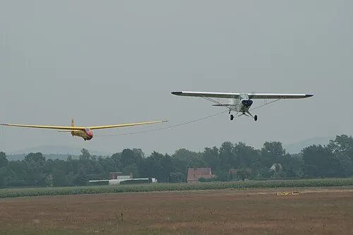 A powered aircraft towing a glider above a grassy airfield.