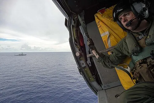 A helmeted person leaning toward the open side of a helicopter above the sea.