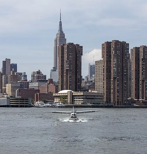 A float-equipped Cessna Caravan on the East River with city buildings behind it.