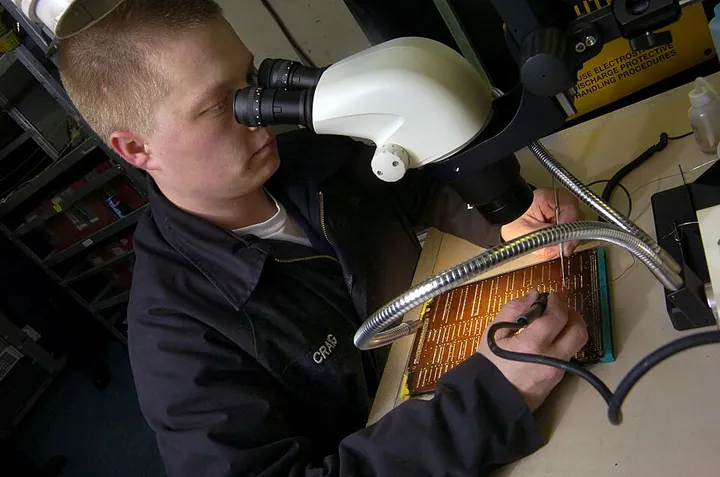 A technician repairs a small circuit card in an avionics workshop.