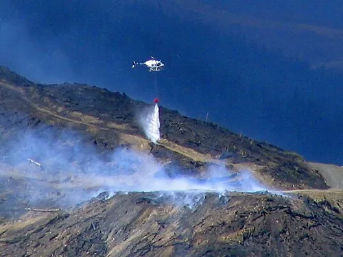 A helicopter releasing water from a suspended bucket over forest.